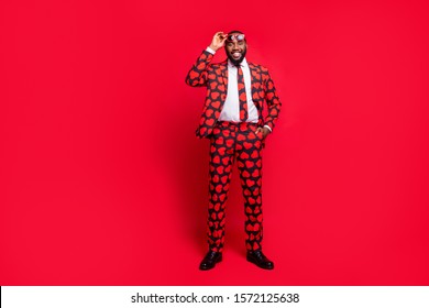 Full body photo of funky dark skin man playing amour cupid character role self-confident wear sun specs hearts pattern suit shirt necktie tie boots outfit isolated red color background