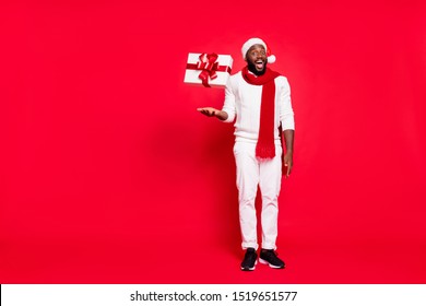 Full Body Photo Of Funny Dark Skin Man Offering Cool X-mas Prices Throwing Giftbox Up Air Amazing Proposition Wear Sweater Trousers Santa Hat Scarf Isolated Red Background