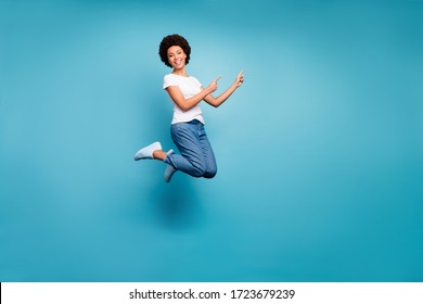 Full body photo of funny dark skin wavy lady jumping high directing fingers empty space offer black friday prices wear casual white t-shirt jeans isolated blue color background