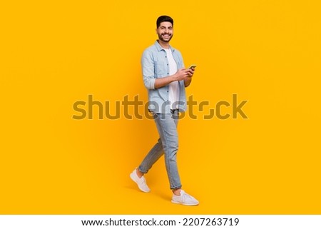 Full body photo of handsome latin young guy model walk hold device read news wear trendy jeans clothes isolated on yellow color background