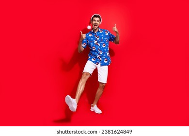 Full body photo of handsome young guy showing double thumb up wear santa claus print x-mas outfit isolated on red color background
