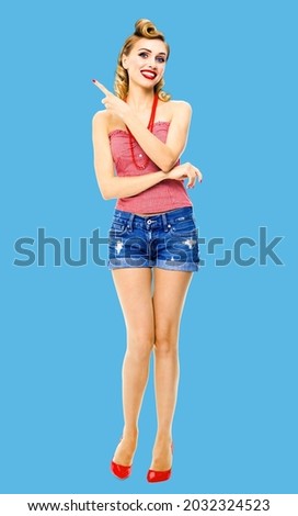 Full body photo - happy smiling woman pointing advertising something. Blond girl in pin up style, showing copy space for ad text or imaginary. Retro fashion and vintage. Blue colour studio background.