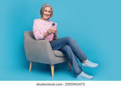 Full body photo of nice senior woman sit armchair hold device wear trendy striped outfit isolated on blue color background - Powered by Shutterstock