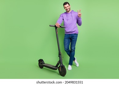 Full body photo of young cheerful man happy positive smile ride scooter show peace v-sign isolated over green color background