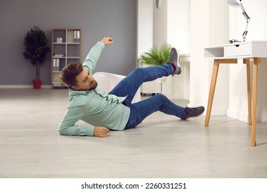 Full body photo of a young clumsy brunette man with unshaven bristles falling down to the floor and injuring his arm at home or in the office at his workplace and wearing casual clothes...
