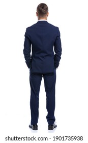 full body picture of back view of young businessman in navy blue suit holding hands in pockets, standing isolated on white background in studio