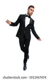 Full Body Picture Of Elegant Young Businessman In Tuxedo Opening Arms, Holding Leg Up And Dancing, Looking To Side On White Background In Studio