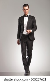 Full Body Picture Of A Young Man In Tuxedo Walking Forward Towards The Camera On Grey Studio Background