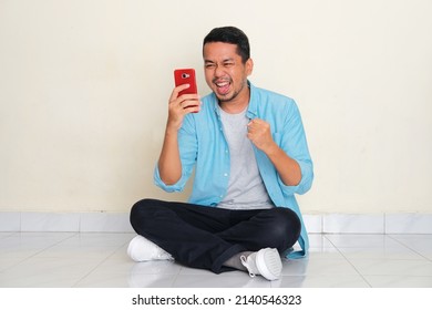 Full Body Portrait Of Adult Asian Man Sitting Cross Legged Showing Winning Gesture When Looking To His Mobile Phone