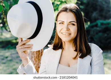 Full Body Portrait Of Caucasian Confident Business Woman In Suit, Walking In Park. Beautiful Woman In Stylish Suit And White Hat Standing Near Tree In The Park