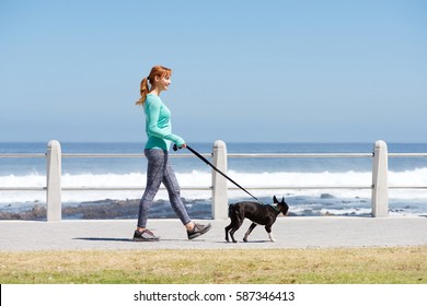 Full Body Portrait Of Fit Woman Smiling And Walking Dog On Path By Sea