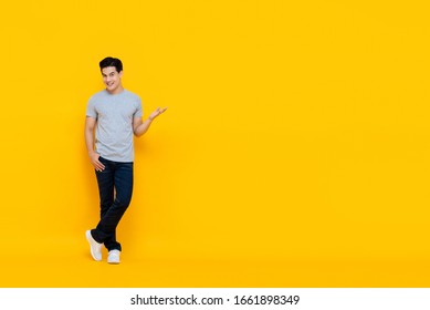 Full body portrait of handsome young Asian man smiling and standing with open hand gesture isolated on yellow background with copy space