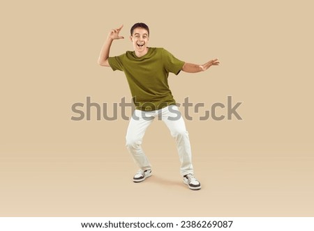 Full body portrait of happy excited cool young funny teenage boy wearing green t-shirt, white jeans and sneakers having fun and dancing isolated on a beige studio background. People emotions concept.