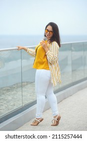 Full Body Portrait Of Happy Millennial Plus Size Woman 30-35 Years Old On Vacation. Smiling Overweight Female In In Yellow And White Fashion Clothes And Sunglasses Drinks Juicy Coctail In Summer Day