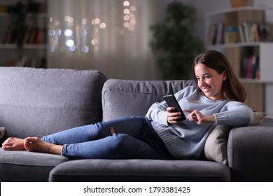 Full Body Portrait Of A Happy Woman Using Smart Phone Lying In The Sofa In The Living Room At Night At Home