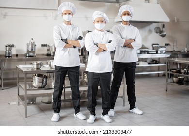 Full body portrait of multiracial team of three chefs standing together in the professional kitchen. Well-dressed chefs in face masks and protective gloves ready for a job. New normal for business - Powered by Shutterstock