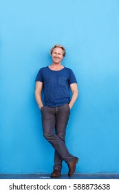 Full Body Portrait Of Older Man Standing Leaning Against Blue Wall And Smiling