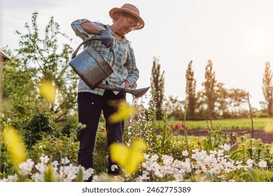 Full body portrait of senior woman watering anemones with watering can in spring garden. Gardener taking care of flowers - Powered by Shutterstock