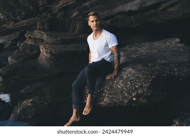 Full body portrait of serious bearded young male in tattooed arm with white t shirt sitting on dark rock of rocky cliff over strea at evening while looking at camera - Powered by Shutterstock