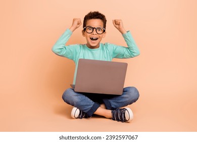 Full body portrait of small delighted schoolchild sit floor use netbook raise fists isolated on beige color background