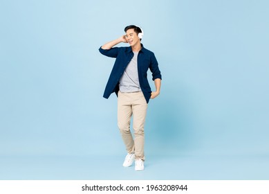 Full Body Portrait Of Smiling Young Handsome Asian Man Listening To Music With Wireless Headphones Isolated On Light Blue Studio Background