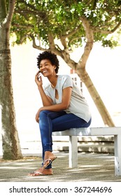 Full Body Portrait Of Smiling Young African Woman Sitting On A Bench Outdoors Talking On Mobile Phone