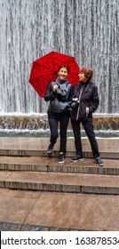 Full Body Portrait Of Two Older Women With Waterfall Fountain In Background In Spring; One Woman Holding Red Umbrella