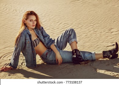 Full Body Portrait Of Young Beautiful Fashionable Woman Posing On The Sand. Model Wearing Blue Denim Jacket, Jeans, Top, Cowboy Boots. Sunset Light. Female Fashion Concept