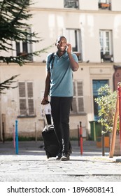 Full body portrait young black man walking with suitcase and talking with cellphone on city street