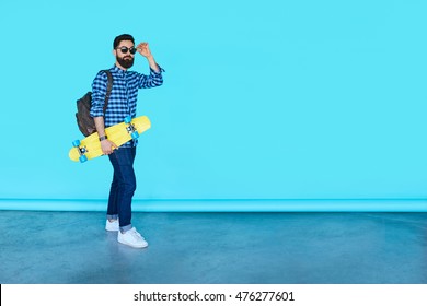 Full Body Portrait Of Young Fashionable Hipster Man Posing Over Blue Background With Copy Space. Bearded Male Holding Yellow Skateboard And Smiling