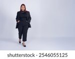 Full body portrait of young Latina businesswoman in black, with red hair and hands in pockets posing taking a step and looking at camera, studio photo with white background and copy space.