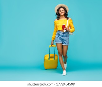 Full body positive ethnic woman with suitcase and tickets smiling and walking towards camera during trip against turquoise backdrop