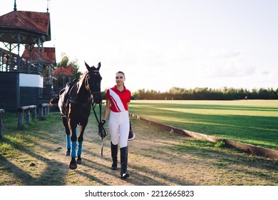 Full Body Of Positive Young Horsewoman With Riding Hat In Hands Holding Horse With Reins And Looking At Camera While Walking On Pathway Near Arena Together In Countryside