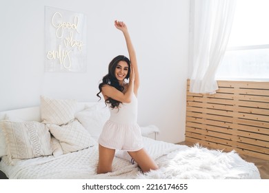 Full Body Of Positive Young Woman With Long Dark Hair Standing On Knees On Comfortable White Bed Near Window And Raising Arm While Looking At Camera In Cozy Bedroom