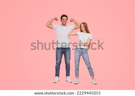 Full body powerful young man and woman in similar clothes smiling and showing biceps against pink background