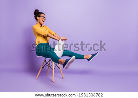Full body profile photo of amazing dark skin lady sitting on chair between legs like driving car wear specs yellow shirt trousers isolated purple color background