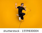 Full body profile photo of crazy hipster guy jumping high holding microphone music lover singing favorite song wear sun specs black t-shirt pants isolated yellow color background