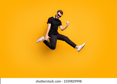 Full body profile photo of crazy hipster guy jumping high holding imagine solo guitar music lover wear sun specs black t-shirt pants isolated yellow color background
