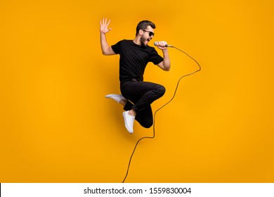 Full body profile photo of crazy hipster guy jumping high holding microphone music lover singing favorite song wear sun specs black t-shirt pants isolated yellow color background