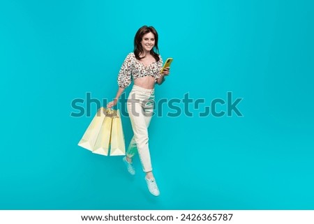 Full body profile portrait of pretty crop top lady use smart phone hold shop bags jump empty space isolated on teal color background
