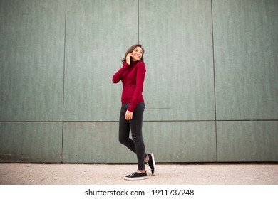 Full body profile portrait young woman walking and talking with cellphone by wall