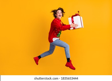 Full body profile side photo of excited girl jump run with x-mas eve noel big gift box, wear christmas tree ornament pullover sweater denim jeans isolated bright shine color background