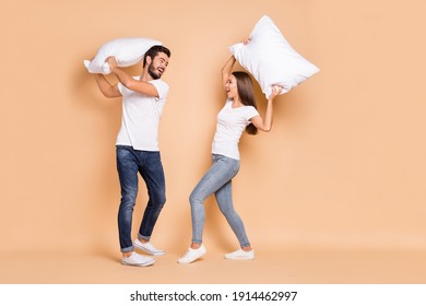 Full body profile side photo of young couple play pillow battle fight happy positive smile excited isolated over beige color background