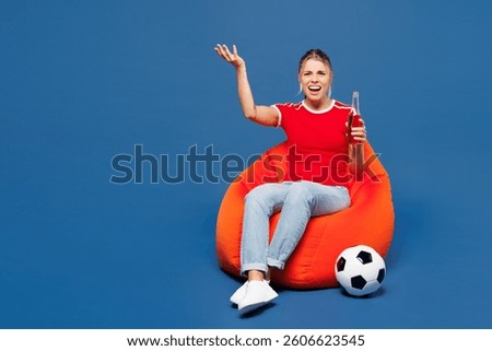 Full body sad mad young woman fan she wear red t-shirt cheer up support football sport team hold in hand soccer ball watch tv live stream sit in bag chair drink beer isolated on plain blue background