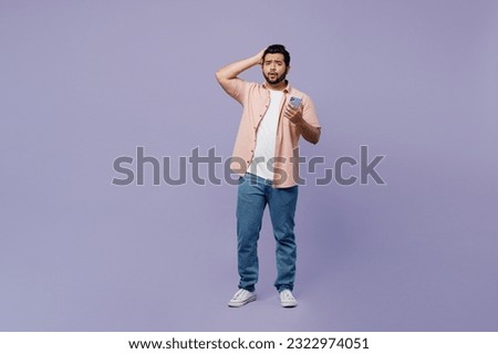 Full body sad shocked scared young Indian man he wear pink shirt white t-shirt casual clothes use mobile cell phone hold head isolated on plain pastel light purple background studio. Lifestyle concept