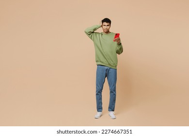 Full body sad young man of African American ethnicity wear green sweatshirt hold scratch head use mobile cell phone isolated on plain pastel light beige background studio . People lifestyle concept - Powered by Shutterstock