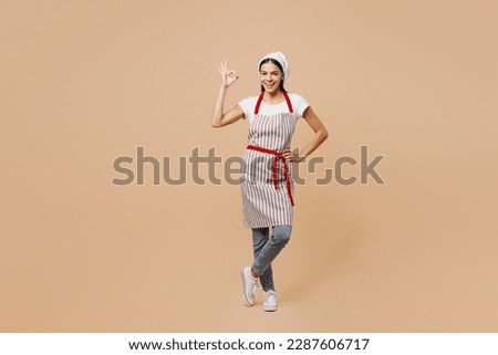 Full body satisfied happy young housewife housekeeper chef baker latin woman wear apron toque hat show ok okay gesture stand akimbo isolated on plain pastel light beige background. Cook food concept