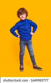 Full body serious kid model in trendy outfit keeping hands on waist and looking at camera against yellow background