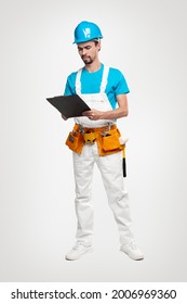 Full Body Of Serious Male Builder In Workwear With Toolkit On Waist Examining Draft On Clipboard While Working On Construction Site Against White Background