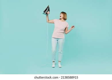 Full Body Shocked Elderly Housewife Woman 50s In Pink T-shirt Gloves Doing Housework Hold In Hand Iron Isolated On Plain Pastel Light Blue Background Studio. Housekeeping Cleaning Tidying Up Concept